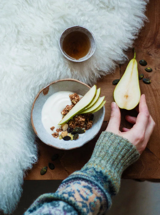 Cozy winter breakfast bowl with Naturally Granola Belgian Dark Chocolate, creamy yogurt, fresh pear slices, and a warm cup of tea, embodying winter self-care and nourishment