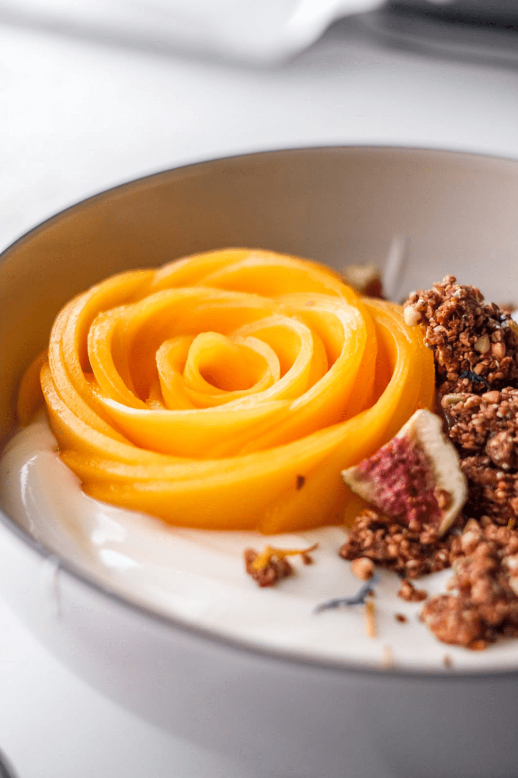 Close-up of a dessert with mango rose, cream, and chunky nute free granola on a white plate.