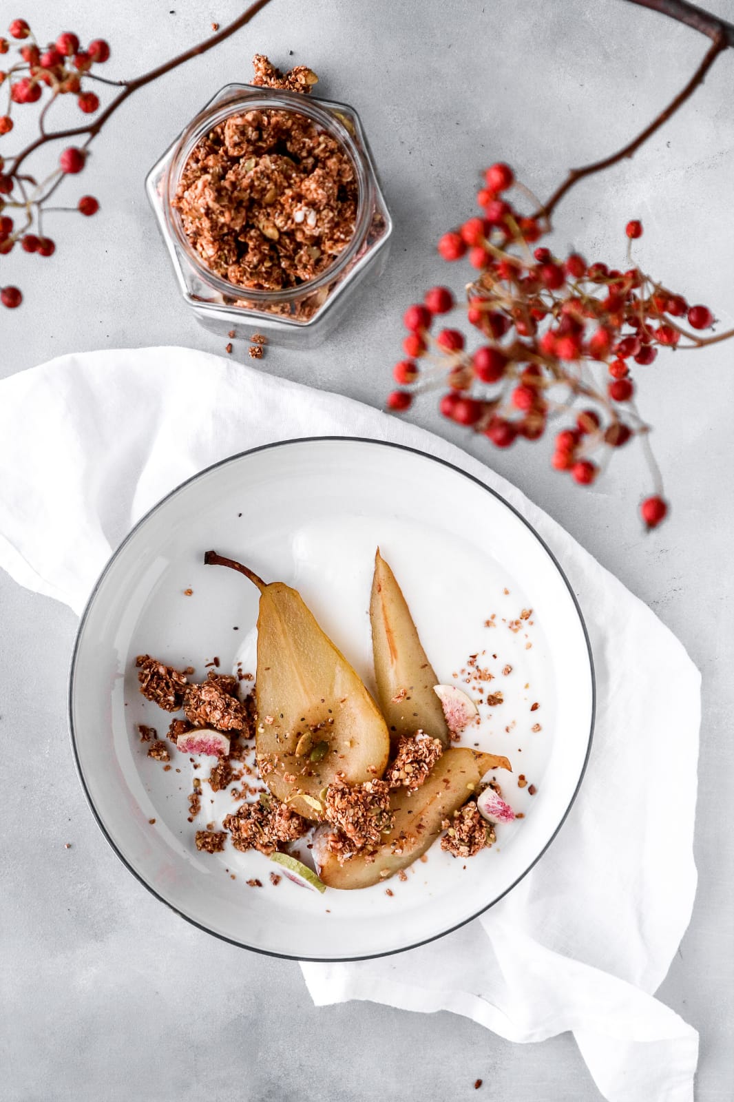Two poached pears on a plate with a chunky granola topping, surrounded by red berries and a small jar of naturally granola on a light gray background.